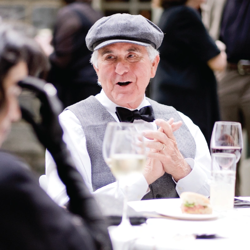 An older man looks to the side, speaking, while sitting at a formally decorated table